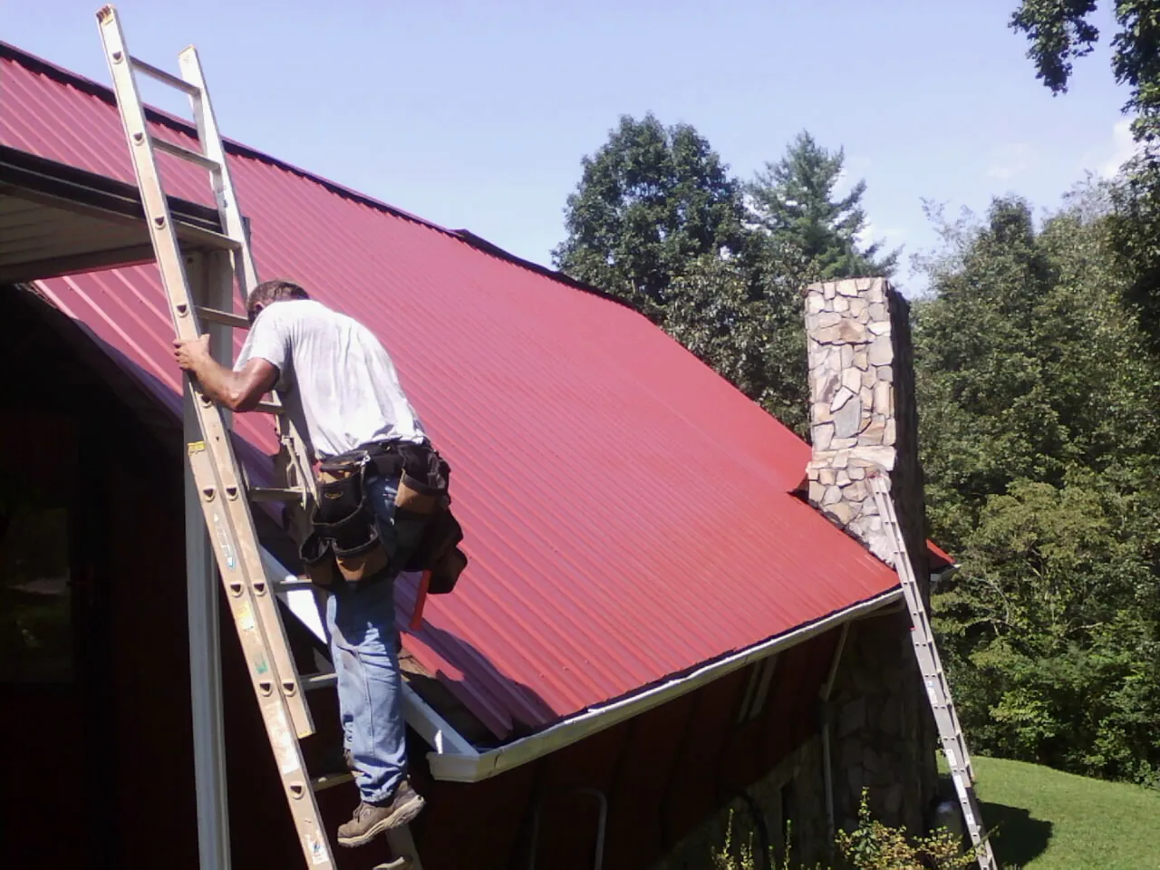 Roofer on a ladder beside a red metal roof and stone chimney — Soffit & Fascia Repair by Roofing Brothers in Marlin